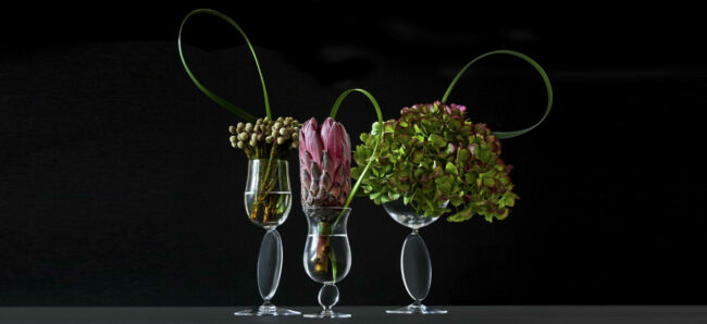 A collection of glasses creating small towers of balance decorated by hydrangeas, leaves and exotic flowers in a black background.